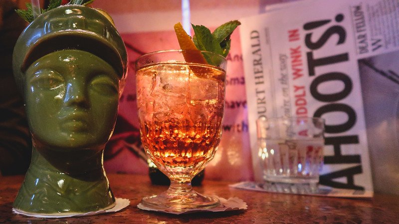 Close-up of cocktails at Cahoots, a 1940s-inspired underground bar in London, featuring a tiki-style green mug and a sparkling drink garnished with mint and orange, set against vintage newspaper décor that enhances the speakeasy’s quirky retro theme