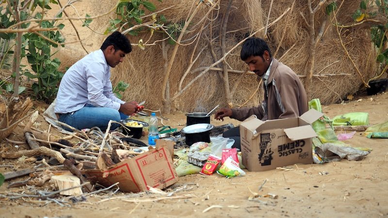 Two local camel safari drivers preparing a traditional meal over an open fire in the Thar Desert near Jaisalmer, India, surrounded by cooking supplies and ingredients on the sand