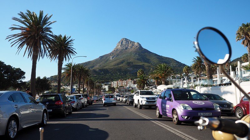 Cruising through Camps Bay, Cape Town on a sidecar motorcycle tour, with a scenic view of Lion’s Head mountain rising in the background, lined palm trees along the bustling coastal road, parked cars, and vibrant city life under a clear blue sky