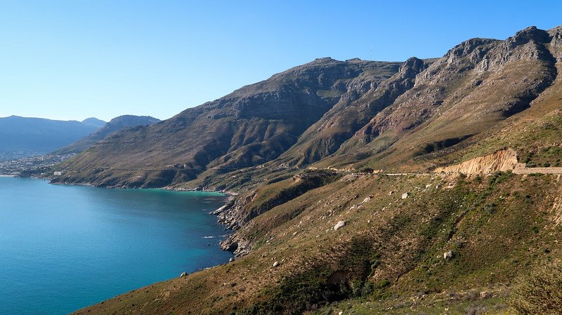 Scenic coastal mountain drive along Camps Bay near Cape Town, South Africa, with rugged cliffs rising above turquoise waters and a winding road hugging the shoreline