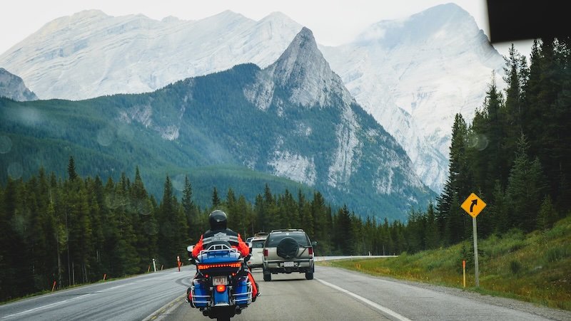 Motorcycle and cars driving along a winding mountain highway surrounded by dense forest and dramatic rocky peaks, capturing the scenic road trip experience through the Canadian Rockies