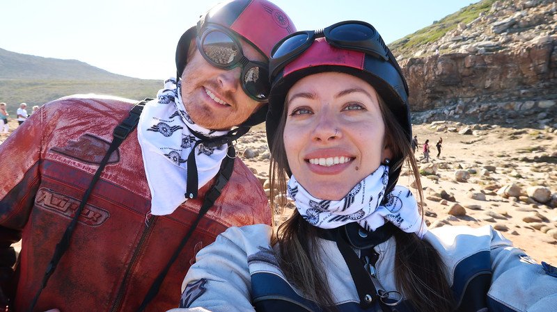 Nomadic Samuel and Audrey Bergner of That Backpacker smiling in a selfie at the Cape of Good Hope during their Cape Peninsula sidecar tour, wearing red and black biker helmets, goggles, and matching riding gear with a rocky coastal landscape behind them