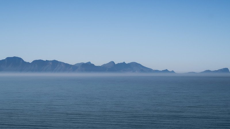 A serene view of the Cape Peninsula coastline with soft morning mist hovering above the water and a distant mountain range silhouetted against the clear blue sky. This peaceful scene was captured during a scenic sidecar tour from Cape Town to Cape Point