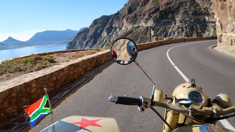 Cruising along the dramatic cliffside roads of the Cape Peninsula in a vintage sidecar motorcycle with Cape Sidecar Adventures, featuring coastal mountain views, the winding road, and a South African flag fluttering in the breeze