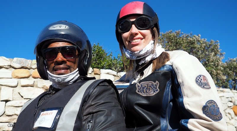 Audrey Bergner of That Backpacker smiling alongside her sidecar driver during a sunny Cape Peninsula Sidecar Tour from Cape Town to Cape of Good Hope, both wearing vintage-style motorcycle helmets and jackets with a bright blue sky overhead