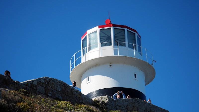 Looking up at the iconic white and black Cape Point Lighthouse with its red roof and panoramic windows, perched on a rocky hill against a clear blue sky, a popular stop during the Cape Peninsula Sidecar Tour from Cape Town to Cape of Good Hope