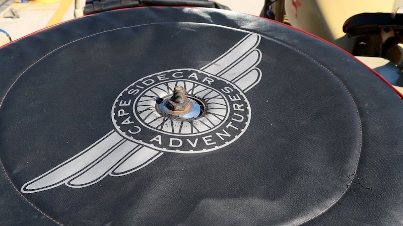 Close-up of the Cape Sidecar Adventures logo printed on the spare tire cover of a vintage sidecar motorcycle, featuring winged graphics and a central wheel emblem, captured during a scenic Cape Peninsula Sidecar Tour from Cape Town to Cape of Good Hope