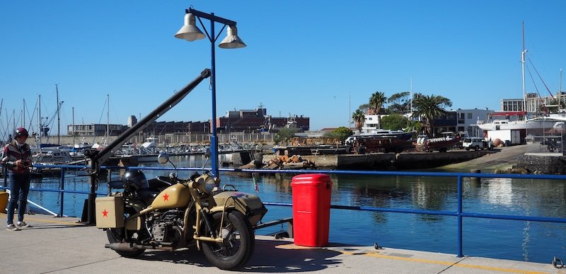 A vintage beige motorcycle with a sidecar featuring red stars is parked along the waterfront under a clear blue sky, with boats and dockside buildings in the background during the Cape Peninsula Sidecar Tour from Cape Town to Cape of Good Hope