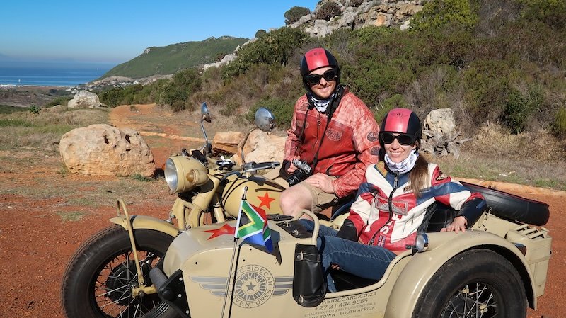Nomadic Samuel and Audrey Bergner of That Backpacker smile while dressed in matching retro biker jackets, helmets, and scarves during their Cape Peninsula Sidecar Tour to the Cape of Good Hope, seated on a vintage motorcycle with scenic coastal views behind