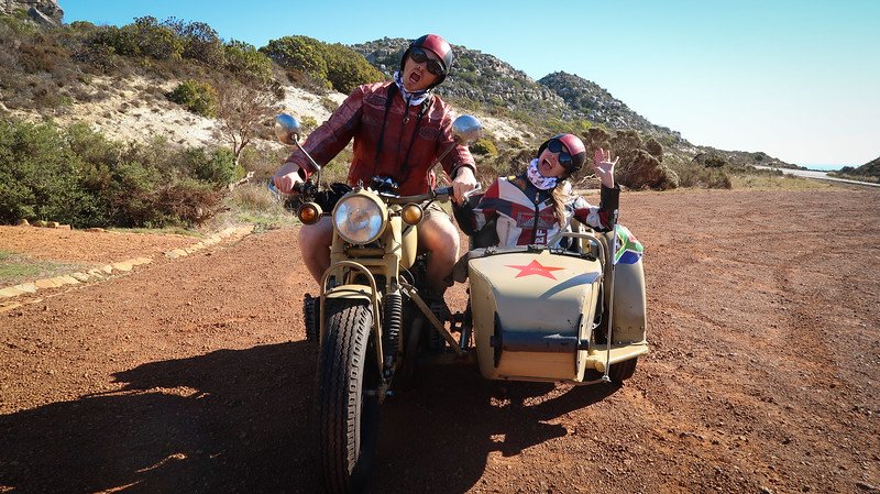 Nomadic Samuel and Audrey Bergner of That Backpacker pose enthusiastically on a vintage motorcycle and sidecar during their Cape Peninsula sidecar tour in South Africa, surrounded by rugged landscapes, red dirt roads, and clear blue skies on a sunny day