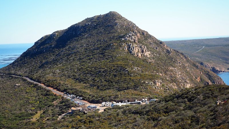 Panoramic view of a rugged, green-covered hill on the Cape Peninsula with a line of tour buses and cars parked at the base, captured during a scenic stop on the Cape Peninsula Sidecar Tour from Cape Town to the Cape of Good Hope, South Africa