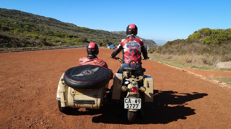 Two riders wearing matching helmets and leather jackets set out on a vintage motorcycle with sidecar during a scenic sidecar tour of the Cape Peninsula near Cape Town, South Africa, with rugged coastal hills in the background