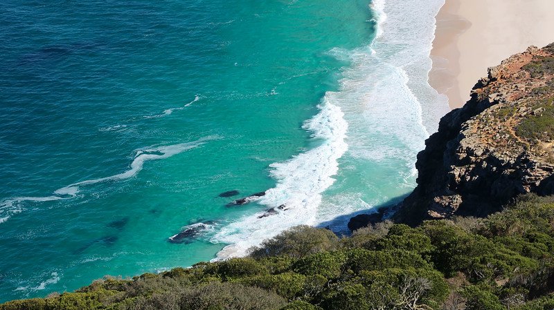 A breathtaking aerial view of turquoise waters and soft sandy shores along the rugged cliffs of Cape Point Nature Reserve in South Africa, showcasing the dramatic coastline and vibrant ocean colors seen during a Cape Peninsula sidecar tour