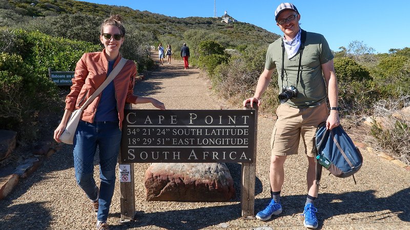 Nomadic Samuel and Audrey Bergner of That Backpacker smiling beside the Cape Point sign in Table Mountain National Park, South Africa, during their Cape Peninsula sidecar tour to the Cape of Good Hope on a sunny day surrounded by coastal vegetation