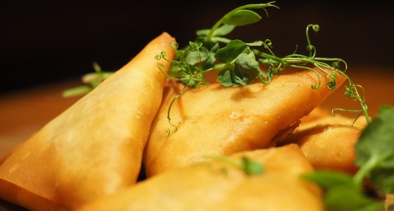 Close-up of golden, crispy samosas garnished with fresh pea shoots, highlighting their flaky texture and rich savory filling — a popular snack to try while visiting Cape Town
