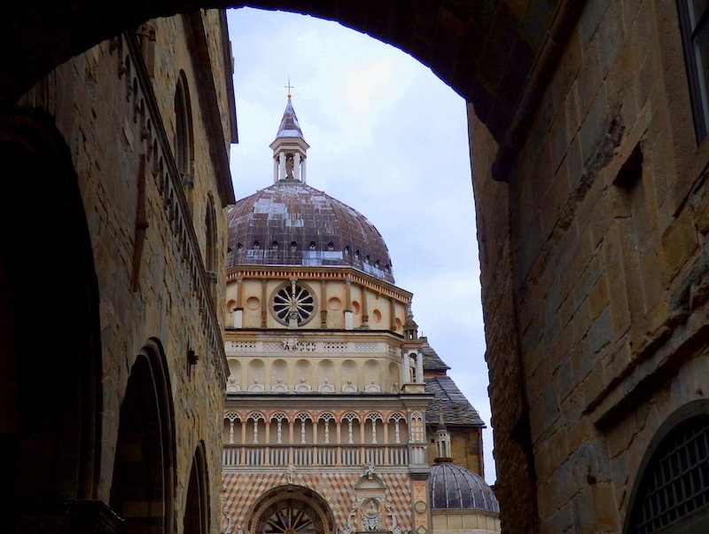 View of the ornate dome and intricate façade of Cappella Colleoni framed by stone archways in Bergamo, Italy, a popular historic day trip from Milan showcasing Renaissance architecture