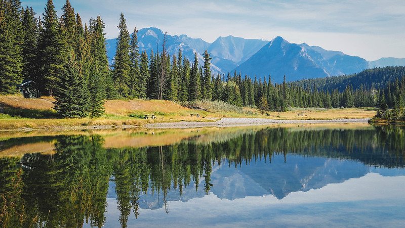 Mirror-like reflections of pine trees and distant rugged peaks on the calm waters of Cascade Ponds, a peaceful picnic spot we visited during our Canadian Rockies family road trip near Banff National Park