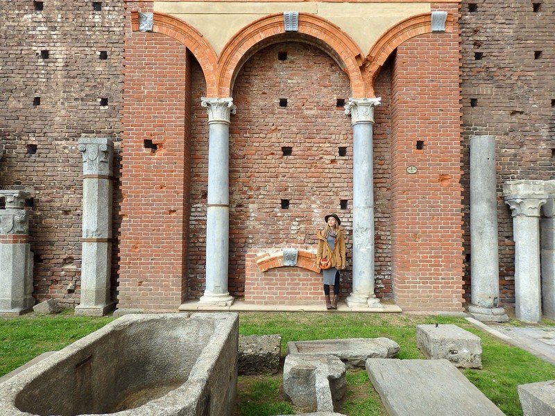Audrey Bergner of That Backpacker standing among ancient stone ruins and marble columns at Castello Sforzesco in Milan, Italy, framed by an old brick archway and Roman-era artifacts