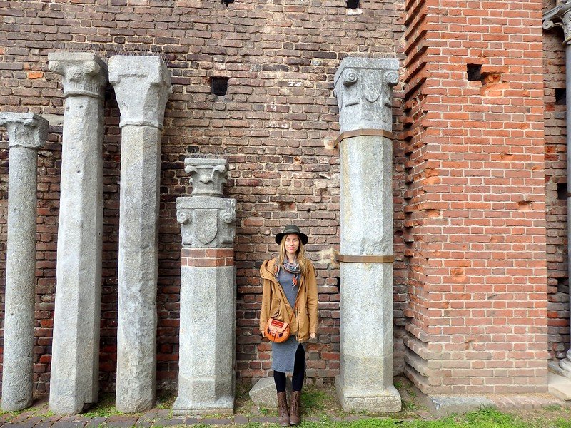 Audrey Bergner from That Backpacker standing among ancient stone columns inside the ruins of Castello Sforzesco in Milan, Italy, showcasing historic architecture and Romanesque details