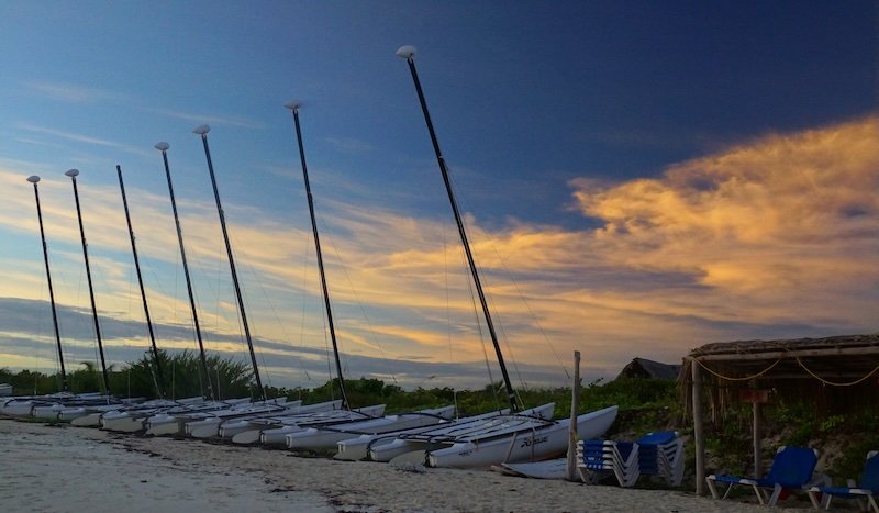 A row of sleek white catamarans with tall masts rests on the sandy shore beneath a golden-hued sunset sky at Cayo Santa María, Cuba, creating a serene and romantic seaside scene during a tropical honeymoon getaway
