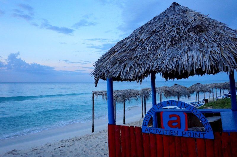 Thatched-roof beach hut marked “Activities Point” overlooking the calm turquoise waters and white sandy shores of Cayo Santa María, Cuba during a peaceful tropical evening