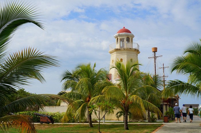 A cream-coloured lighthouse with a red dome rises above swaying palm trees under partly cloudy skies, capturing the tropical scenery and coastal charm of Cayo Santa María, Cuba during a sunny beach getaway