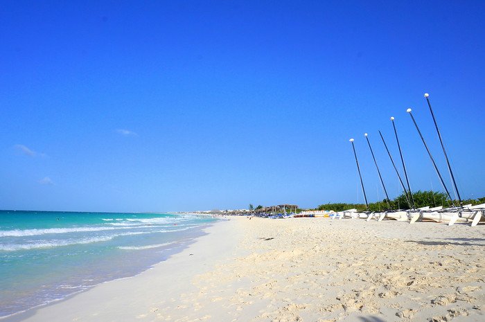 A pristine white sand beach with gentle turquoise waves and sailboats lined up along the shore under a cloudless blue sky in Cayo Santa María, Cuba, captured during a sunny and relaxing honeymoon getaway