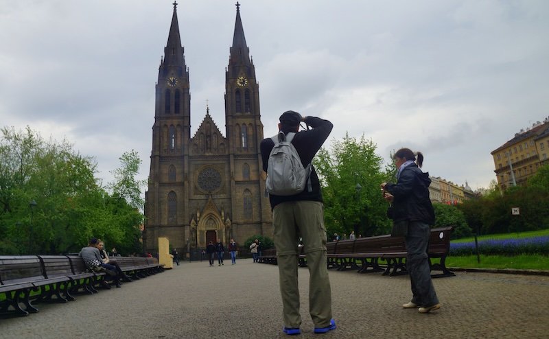 Visitors taking photos of the striking neo-Gothic Church of St. Ludmila with its twin spires rising above Peace Square in Prague, Czech Republic, on a cloudy afternoon