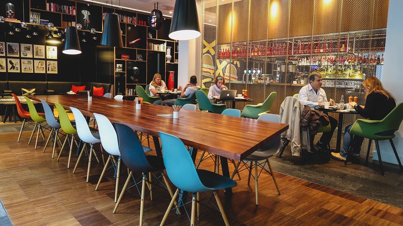 Guests enjoying breakfast and working on laptops inside the stylish CitizenM Hotel in London, featuring a long wooden communal table, colorful modern chairs, and a vibrant atmosphere that blends dining, work, and social space seamlessly