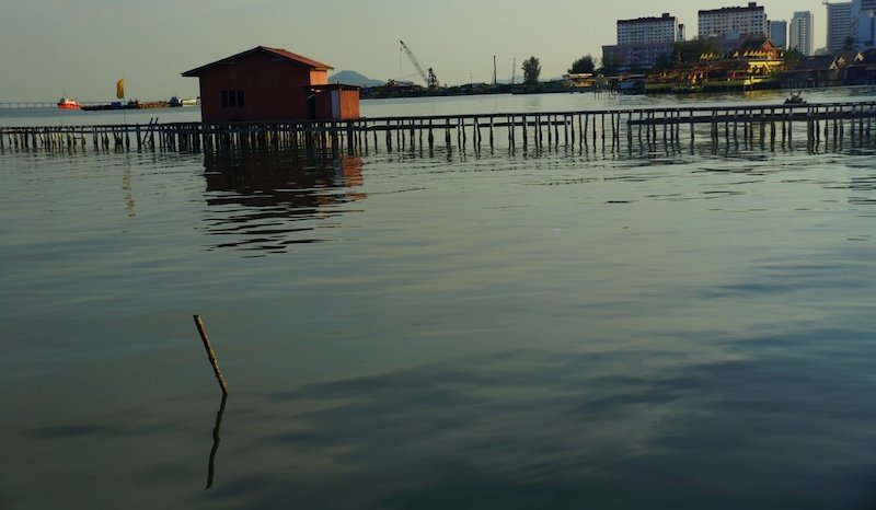 View of a traditional wooden stilt house at the Clan Jetties in Georgetown, Penang, Malaysia, with long pier stretching over the water and city skyline in the background