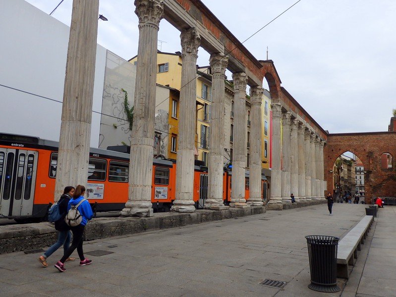 Ancient Roman columns of Colonne di San Lorenzo standing along a bustling street in Milan, Italy, with orange trams passing behind and pedestrians walking by this historic landmark