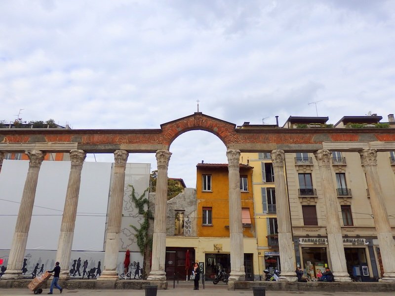 Ancient Roman columns of Colonne di San Lorenzo framed by colorful residential buildings in Milan, Italy, blending historic architecture with modern city life