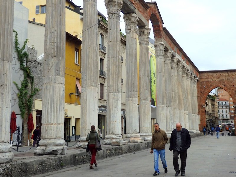 Visitors strolling past the ancient Roman columns of Colonne di San Lorenzo in Milan, Italy, surrounded by historic architecture blending with modern city life