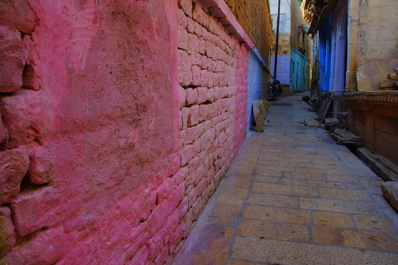 A narrow alleyway in Jaisalmer, Rajasthan, India, featuring vibrant pink, blue, and purple painted walls contrasting with the golden sandstone architecture of the old city