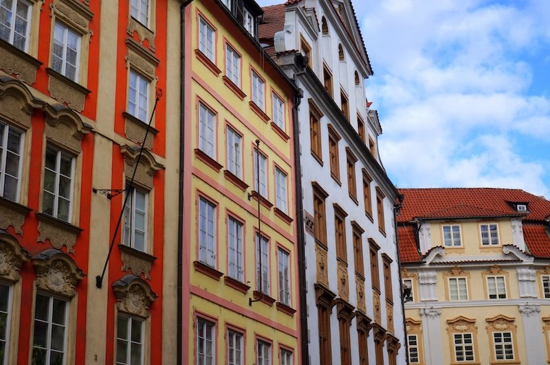 Row of vibrant baroque and rococo-style buildings painted in orange, yellow, and white with ornate facades and red-tiled roofs in Old Town Prague, Czech Republic, under a bright blue sky.