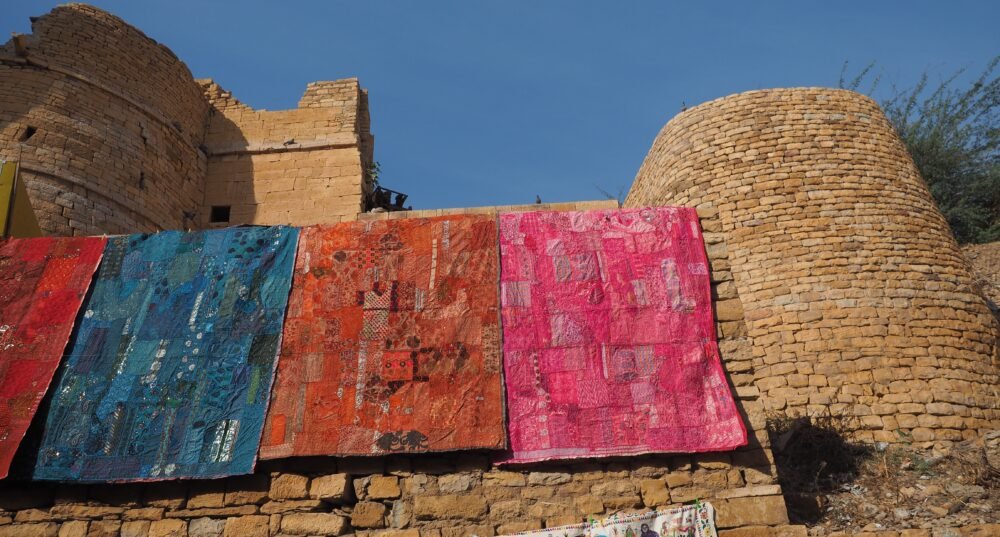 Vibrant handmade blankets in rich blue, orange, and pink hues hanging along the ancient golden sandstone walls of Jaisalmer Fort in Jaisalmer, Rajasthan, India, creating a colorful contrast against the historic desert fortress