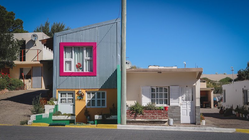 Two colourful neighbouring homes on Avenida de las Ballenas in Puerto Pirámides, Patagonia, Argentina, stand side by side under a bright blue sky. The playful architecture and vibrant accents reflect the relaxed coastal village charm of this destination