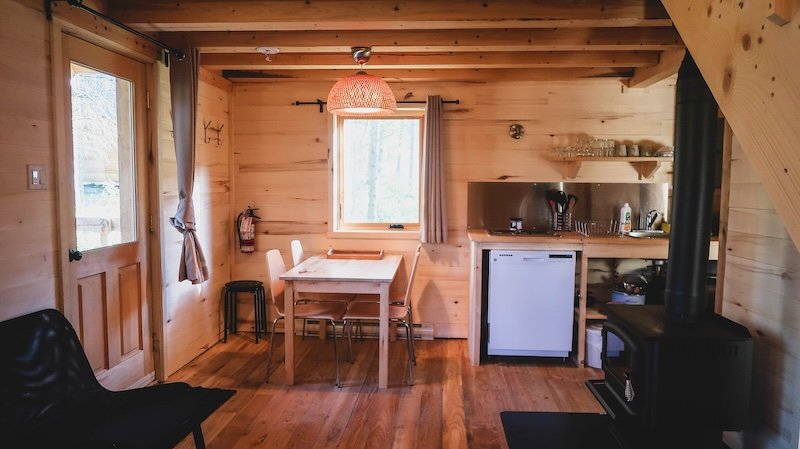 Cozy wooden cabin interior at Sutton in Quebec’s Eastern Townships, featuring a warm wood-panelled design, dining table, compact kitchen, and wood-burning stove