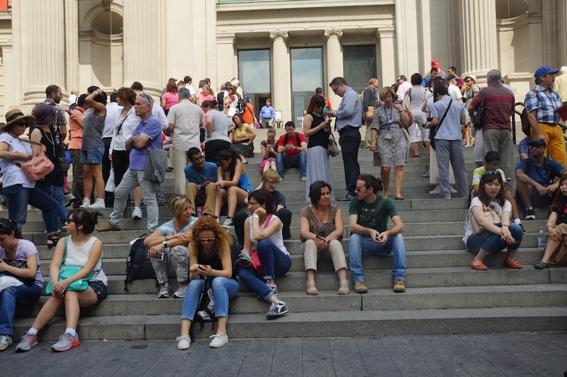 A large crowd of visitors sits and gathers on the famous steps of the Metropolitan Museum of Art in New York City on a sunny day, capturing the vibrant energy of tourists and locals enjoying one of the city’s most iconic cultural landmarks
