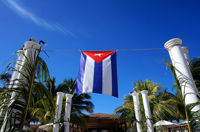 The Cuba national flag flutters brightly against a vivid blue sky, framed by tall white columns and swaying palm trees at a tropical resort in Cayo Santa María during a romantic honeymoon getaway