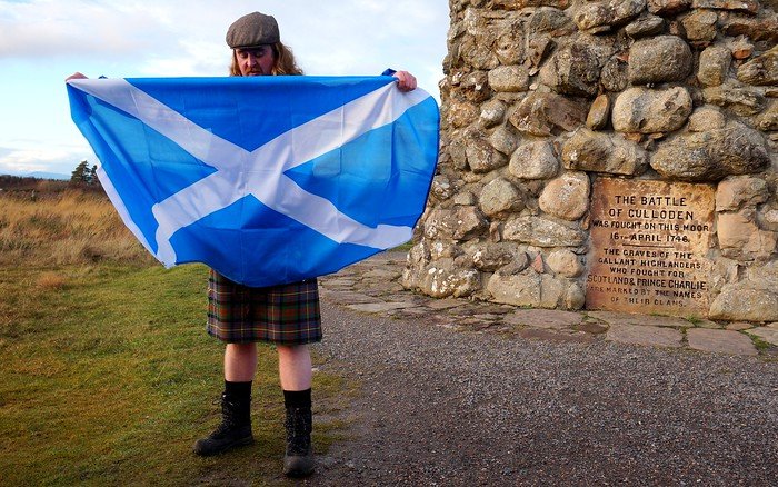 Tour guide in a kilt holding the Scottish flag at Culloden Battlefield in Scotland, standing near the stone memorial that marks the site of the 1746 battle, sharing the history of the Jacobite uprising and its lasting significance