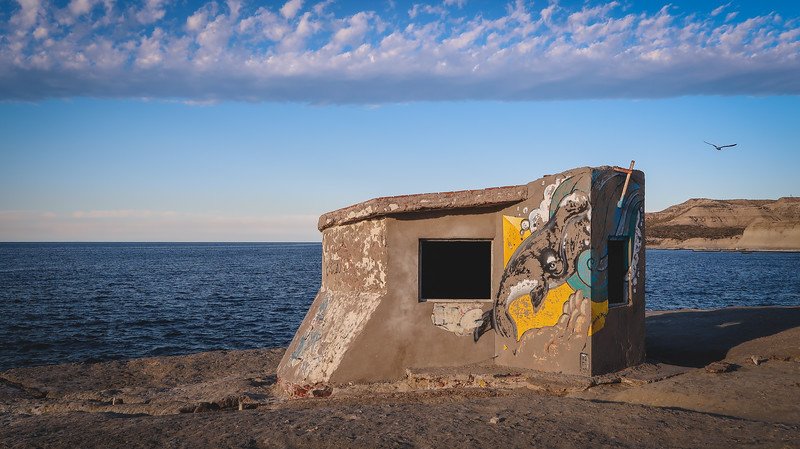 A weathered concrete structure with a vibrant dolphin mural stands on the cliffs overlooking the Atlantic Ocean in Puerto Pirámides, Península Valdés. The golden light and dramatic sky create a striking scene along the coastal walking trail.