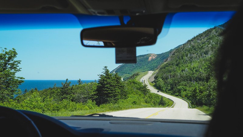 View from inside a car driving along the scenic Cabot Trail in Cape Breton Island, Nova Scotia, with a winding coastal road cutting through lush green forested hills and glimpses of the blue Atlantic Ocean ahead