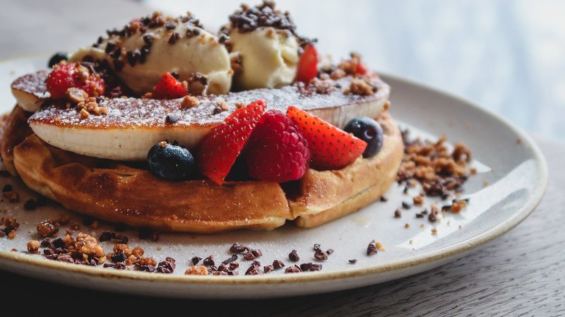 Close-up of a decadent dessert waffle at Duck & Waffle London, topped with caramelized banana, scoops of vanilla ice cream, fresh berries including strawberries, raspberries, and blueberries, and sprinkled with crunchy granola and cocoa nibs