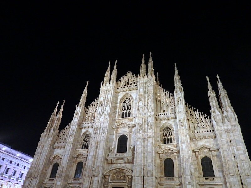 Illuminated façade of Milan’s iconic Duomo di Milano at night, showcasing its intricate Gothic spires and detailed marble architecture against a dark sky.