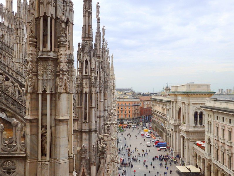 Panoramic rooftop view from Milan’s iconic Duomo di Milano, showcasing its intricate Gothic spires overlooking the bustling Piazza del Duomo and surrounding historic architecture