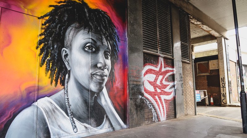 Striking street art mural in East Croydon, London, depicting a detailed black-and-white portrait of a woman with braided hair against a vivid gradient background of orange, pink, and purple, showcasing the neighborhood’s vibrant urban art culture