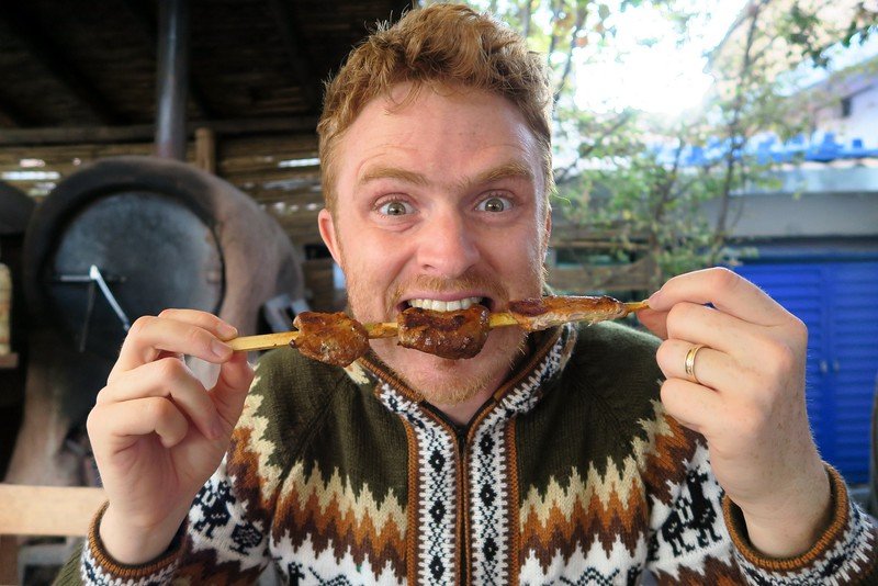 Eating alpaca skewers in Cuzco, Peru as traditional Peruvian cuisine is a hungry Nomadic Samuel eagerly taking a bit bite of the food