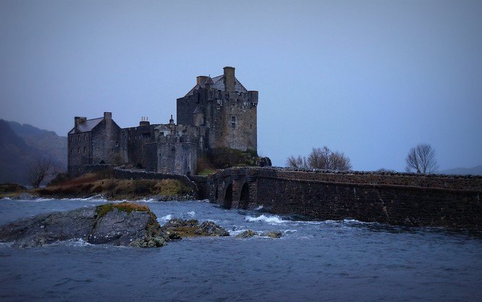 Eilean Donan Castle in the Scottish Highlands, set on a small island where three lochs meet, connected by a stone bridge, with waves splashing along the shore under a misty sky, creating a dramatic and iconic Scotland landscape
