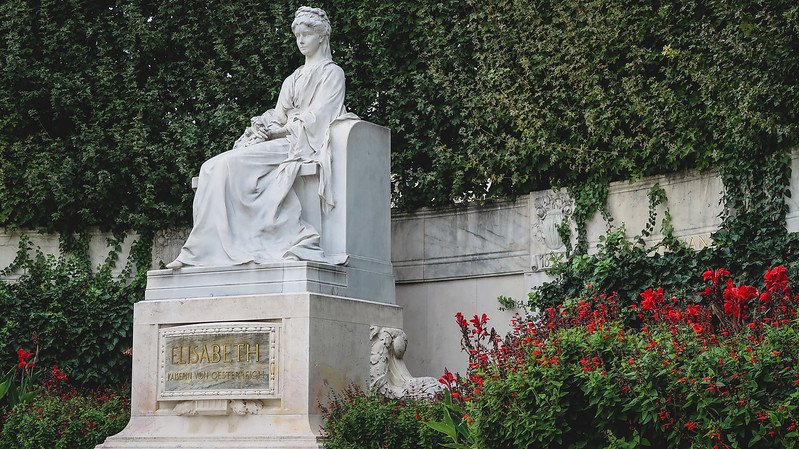 Elegant white marble statue of Empress Elisabeth of Austria (Sisi) surrounded by vibrant red flowers and lush greenery in Vienna, Austria Elegant white marble statue of Empress Elisabeth of Austria (Sisi) surrounded by vibrant red flowers and lush greenery in Vienna, Austria